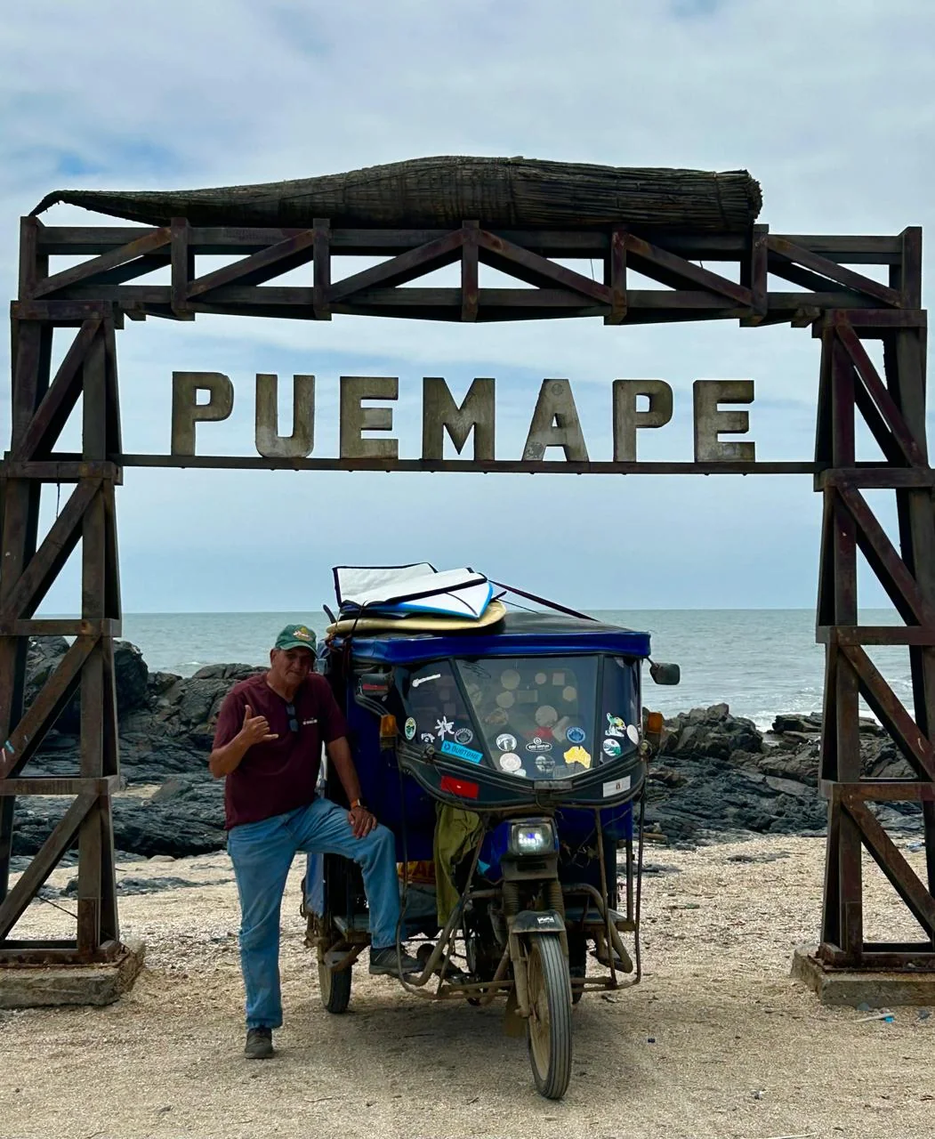 Tato con su mototaxi y las tablas bajo el arco de entrada de Pu&eacute;mape